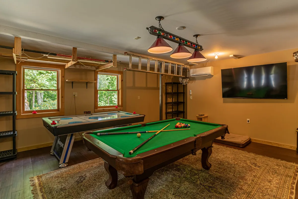 This image features a well-lit, inviting game room with a pool table and air hockey table as the central attractions. A wall-mounted TV and windows provide both entertainment and natural light, while the open space allows for group interaction and playful competition.