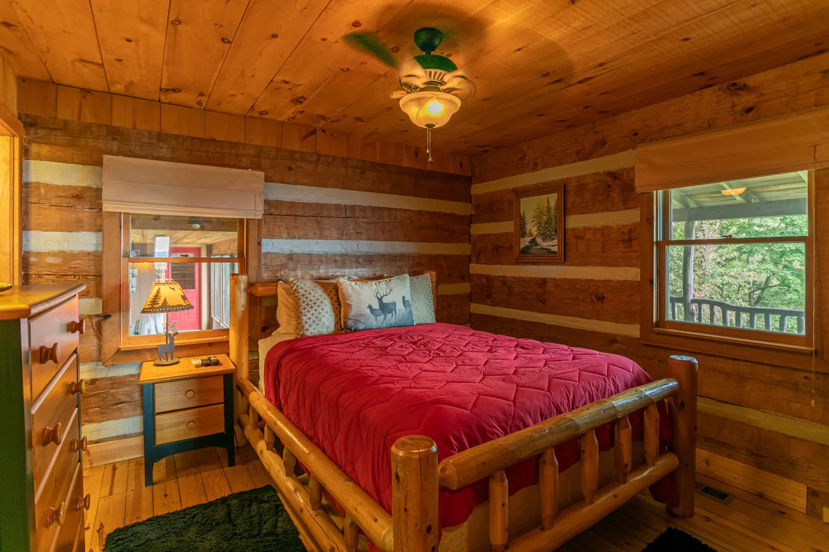 A cozy bedroom featuring a rustic log bed topped with a red quilt, set against warm wooden walls. A window offers a view of the surrounding nature, filling the room with soft daylight and inviting a sense of calm. The space feels intimate and welcoming, ideal for restful retreats.