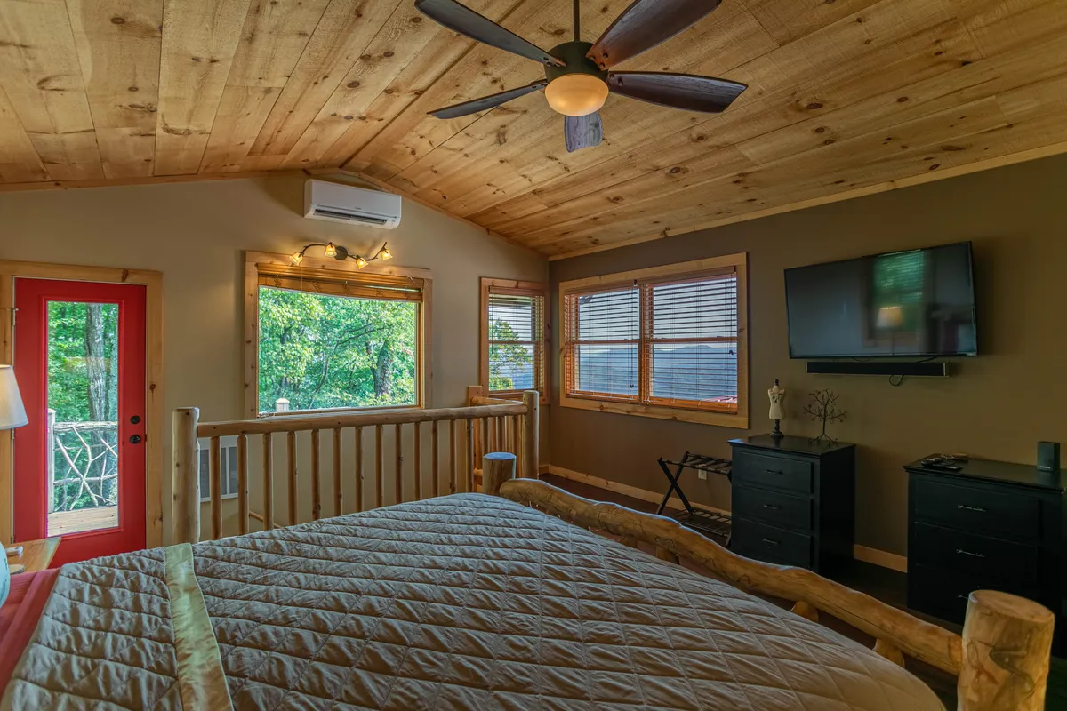 A spacious master suite boasting a king-sized bed with a quilted cover, a flat-screen TV, and large nightstands. A red door leads outside, hinting at private outdoor access. Wooden ceilings and walls add to the serene, luxurious feel, making it a true sanctuary for couples or anyone seeking privacy.