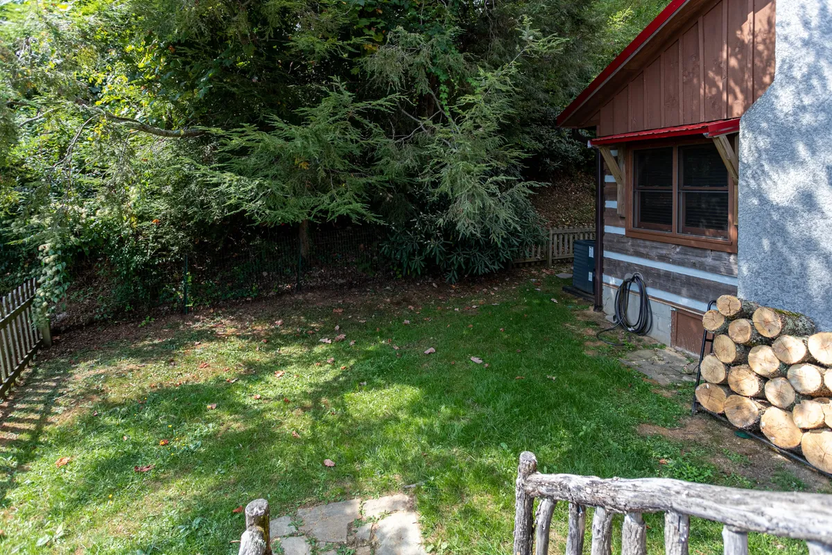 The exterior yard of the cabin, featuring a log pile, wooden fence, grassy area, and mature tree canopy. The log cabin structure is visible, giving a sense of the natural, inviting environment perfect for group gatherings and outdoor enjoyment.
