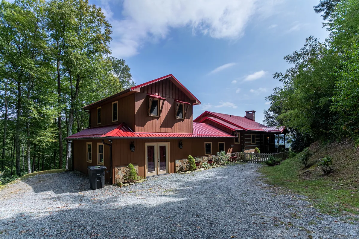 The cabin’s charming exterior features a red roof and wooden facade nestled among lush trees. The gravel driveway suggests easy access and a welcoming arrival, while the surrounding woods invite exploration. The image hints at nearby hiking trails and outdoor gathering spaces.