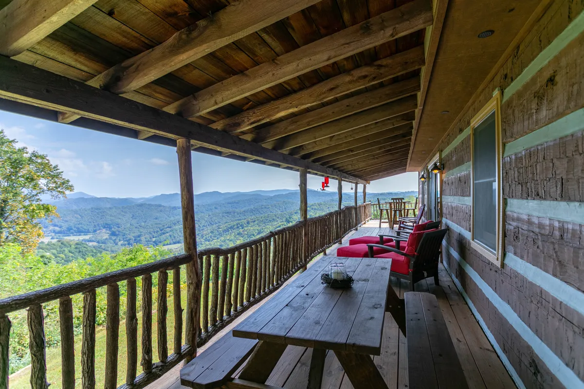 A spacious wooden deck with a rustic railing overlooks a sweeping mountain view. Red chairs and a long wooden table invite guests to dine outdoors or simply relax in the fresh air. The deck is surrounded by the changing colors of the trees, emphasizing its year-round appeal for gatherings and scenic appreciation.