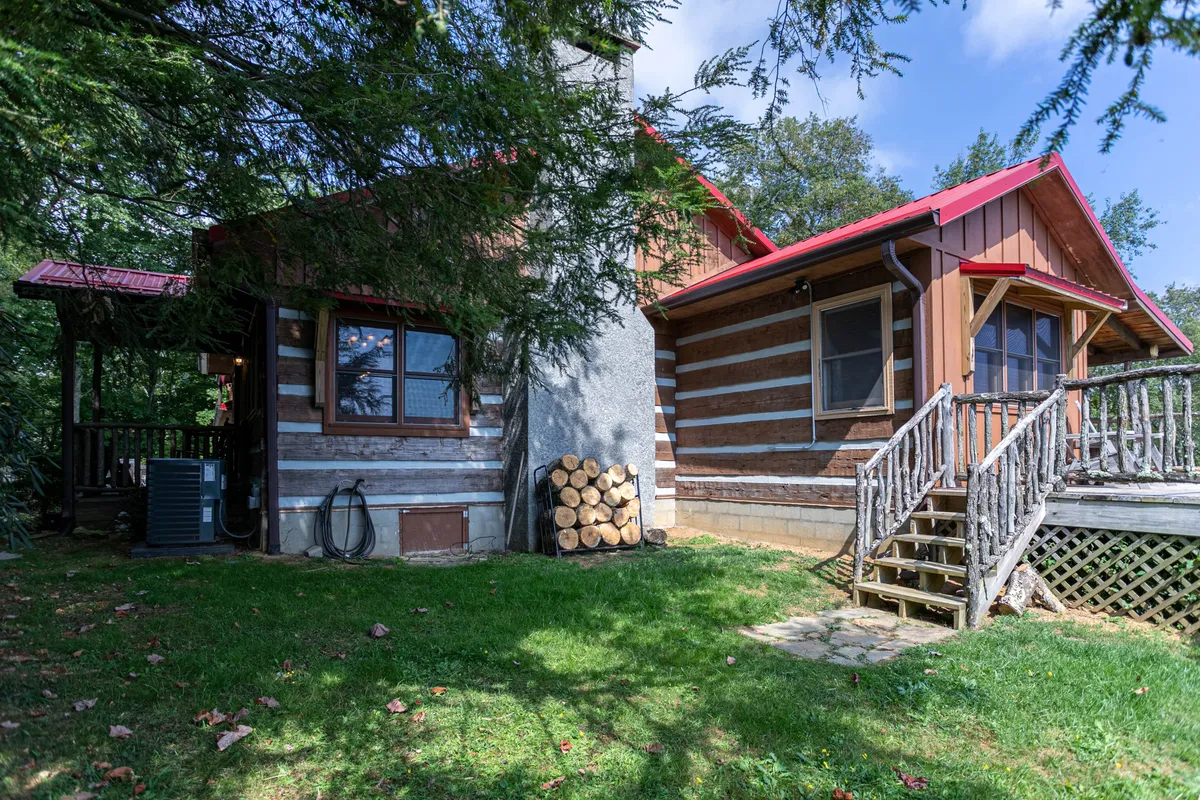 This exterior shot features the log cabin structure, wooden steps leading up to the porch, and a visible stack of firewood. The foreground shows a welcoming porch area ideal for relaxing or letting pets explore a contained area before heading out to trails. The surrounding trees and rustic details evoke a sense of natural mountain living.