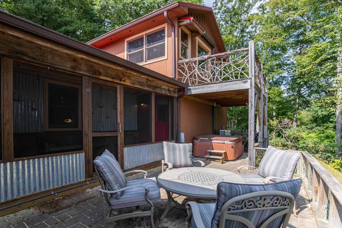 A wooden deck with an outdoor seating area and hot tub, bordered by a rustic railing with natural patterns. The space is surrounded by lush nature, offering opportunities for relaxation and group entertainment.