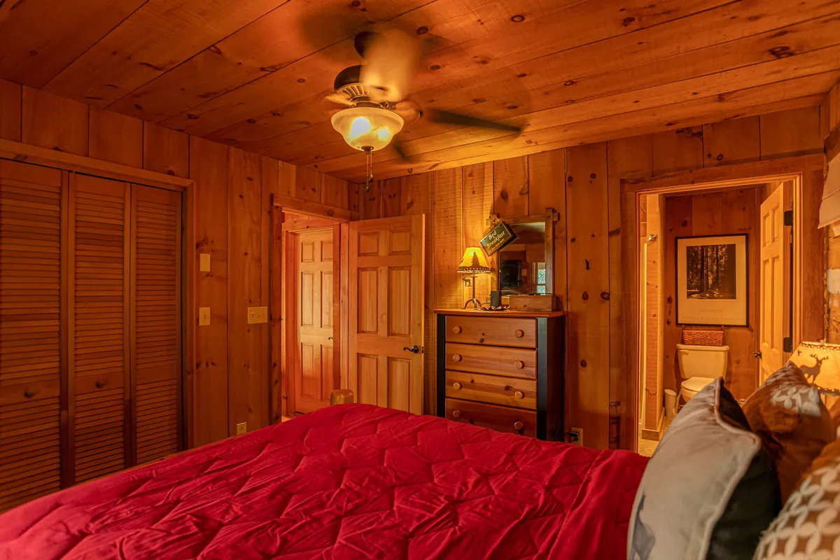 A warm, inviting bedroom with a red quilted bedspread, a wooden dresser, ceiling fan, lamp, and rich wooden paneling. The lighting is soft, and the room feels enveloping—perfect for quiet nights and relaxation after a day in the mountains.