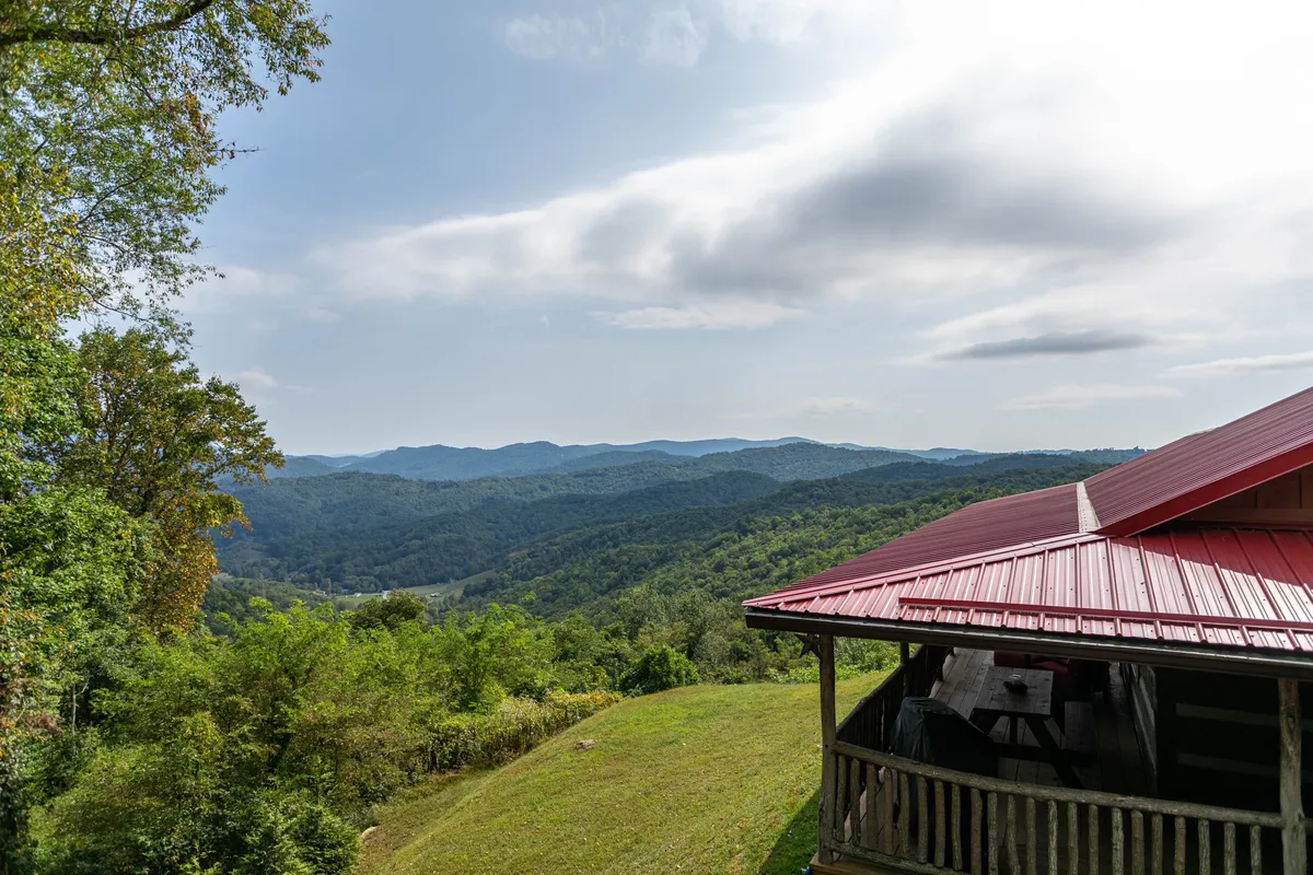 The image showcases a classic red-roofed log cabin perched atop a lush, green mountaintop, with dramatic, sweeping views of the Blue Ridge Mountains stretching into the distance. The sunset bathes the landscape in golden light, illuminating the cabin’s inviting porch and cozy fire pit area, with wildflowers dotting the foreground.