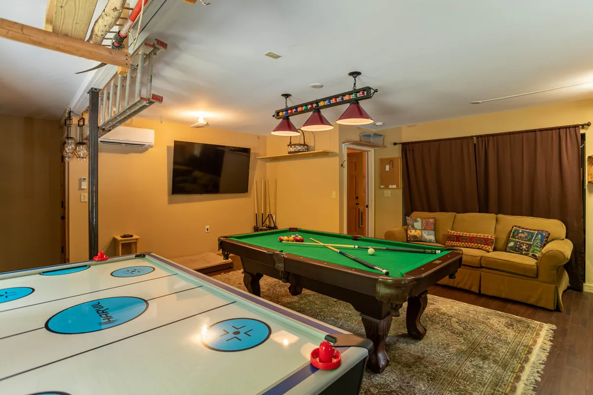 A vibrant game room setting with both a pool table and an air hockey table. A comfortable sofa faces a large TV, suggesting a multifunctional space for both active games and relaxed viewing. The arrangement encourages socializing and friendly competition.