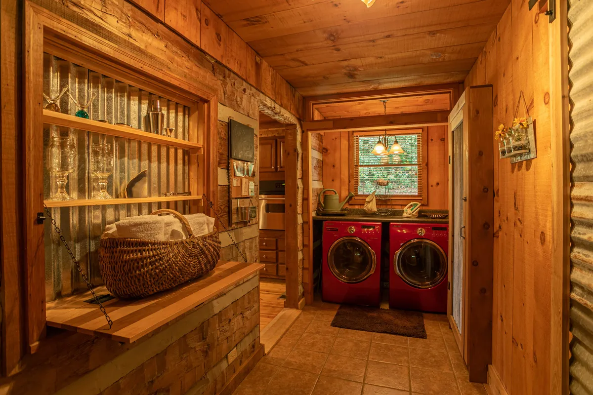 A practical, inviting nook with high-end red washing machines, wooden shelving stocked with glassware and linens, and a window overlooking greenery. The space feels both functional and luxurious.